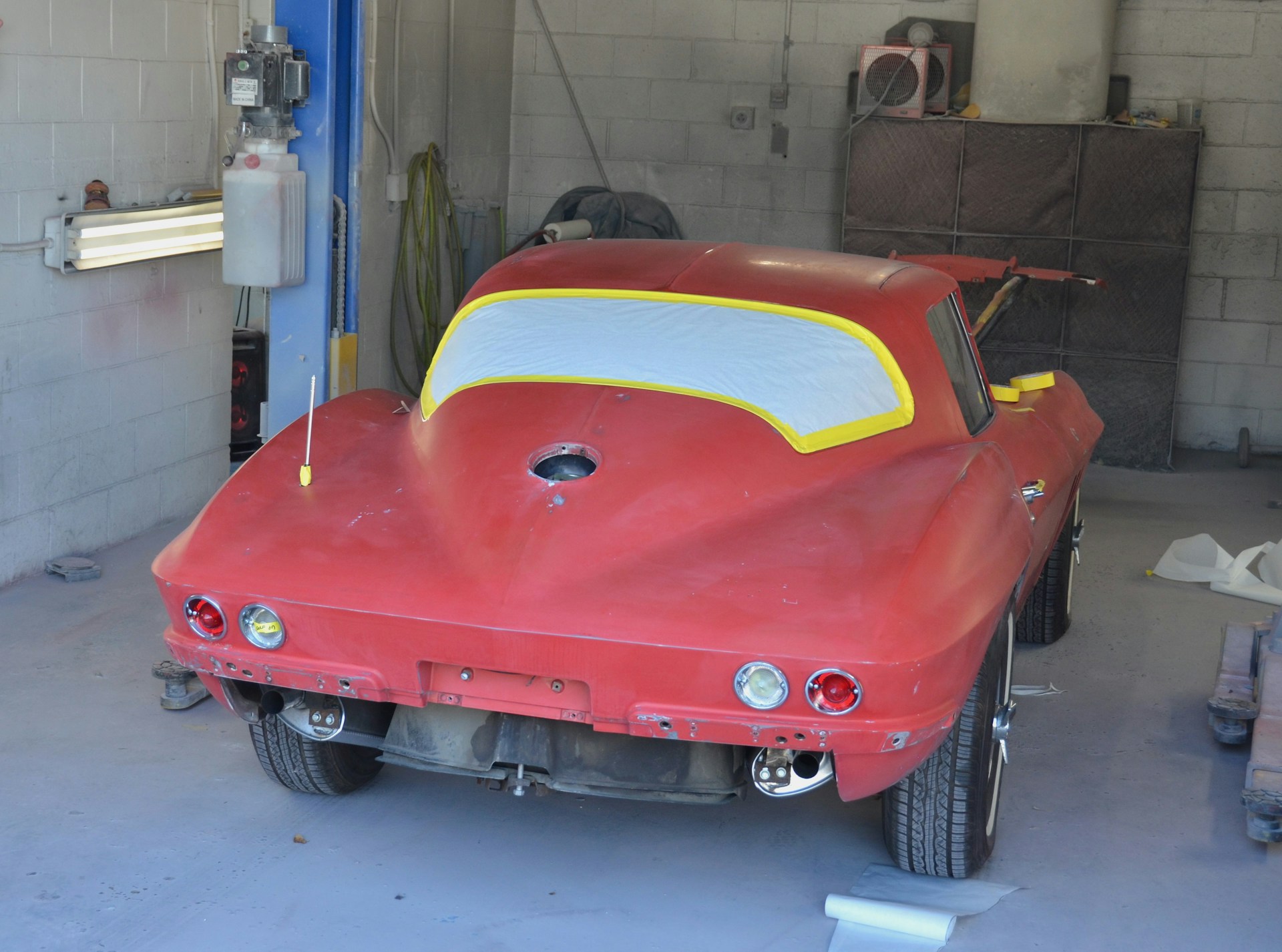 A partially restored or prepared vintage red car is inside a garage, with masking paper covering the windows and some unfinished or missing parts. The setting includes a concrete wall and typical garage equipment.