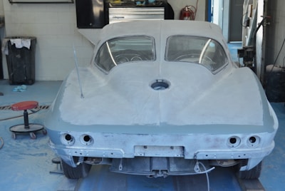 The image features a partially restored vintage car in a garage or workshop. The car is covered in a grey primer and appears to be a classic model with distinctive rear curves and round tail lights. The workshop environment includes a mechanic's stool, tools, and other equipment visible around the car.