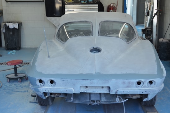 The image features a partially restored vintage car in a garage or workshop. The car is covered in a grey primer and appears to be a classic model with distinctive rear curves and round tail lights. The workshop environment includes a mechanic's stool, tools, and other equipment visible around the car.