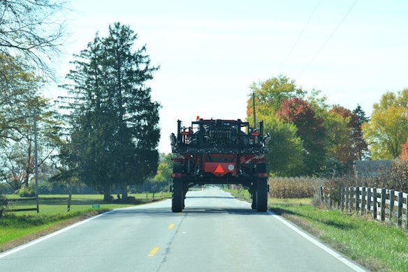 A large agricultural vehicle is traveling down a rural road. The road is flanked by trees displaying autumn foliage with green, yellow, and red leaves. The sky is clear and bright, and the setting exudes a calm and serene countryside vibe.