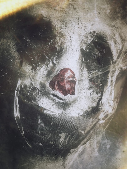 A haunting and abstract portrayal of a face-like figure emerges through a textured, translucent material. The rough surface appears scratched and worn, while a dark central element resembles a nose, adding a sense of mystery and unease.