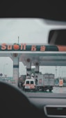 A gas station with multiple fueling pumps and several vehicles parked. Signs and logos in different languages are visible, and the setting appears overcast. Vehicles include a large truck and a white van.