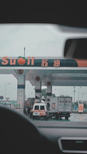 A gas station with multiple fueling pumps and several vehicles parked. Signs and logos in different languages are visible, and the setting appears overcast. Vehicles include a large truck and a white van.