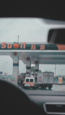 A gas station with multiple fueling pumps and several vehicles parked. Signs and logos in different languages are visible, and the setting appears overcast. Vehicles include a large truck and a white van.