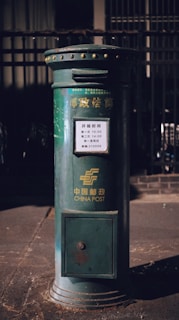 A vintage-style green postbox is prominently positioned on the street, bearing the logo and text of China Post. The postbox includes a schedule in Chinese characters with collection times listed.