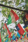 A collection of colorful international flags hangs on strings amidst lush green foliage. The leaves create a natural backdrop, with sunlight filtering through, contributing to a vibrant and lively atmosphere. Various flags represent different countries, indicating multicultural diversity.