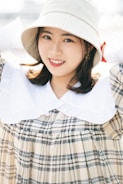 A person wearing a wide-brimmed hat and a checked dress with a white collar. The individual has a warm, friendly smile and the background is softly blurred, suggesting a bright, sunny environment.
