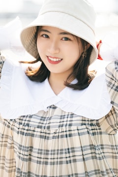 A person wearing a wide-brimmed hat and a checked dress with a white collar. The individual has a warm, friendly smile and the background is softly blurred, suggesting a bright, sunny environment.