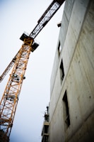 A towering construction crane next to a partially built concrete structure. The crane is orange with an intricate lattice framework. The building appears to be in the initial stages of development with visible scaffolding and open window spaces.