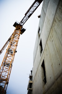 A towering construction crane next to a partially built concrete structure. The crane is orange with an intricate lattice framework. The building appears to be in the initial stages of development with visible scaffolding and open window spaces.