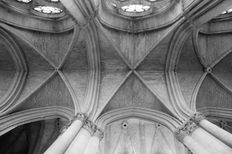Intricate stone arches and vaulted ceilings form a complex geometric pattern. The architecture is characterized by detailed masonry work and grand columns. Light filters through circular stained glass windows, adding depth and elegance to the structure.