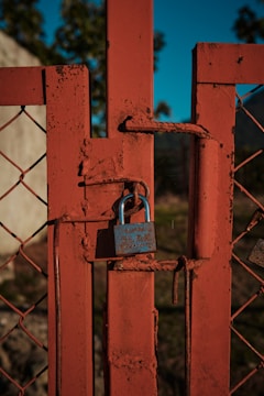 A rusty metal gate is secured with a blue padlock. The gate features a diamond-shaped wire mesh and is painted in a faded reddish-orange color. The background includes a hint of blurred greenery and a wall, indicating an outdoor setting likely in a rural or industrial area.