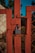 A rusty metal gate is secured with a blue padlock. The gate features a diamond-shaped wire mesh and is painted in a faded reddish-orange color. The background includes a hint of blurred greenery and a wall, indicating an outdoor setting likely in a rural or industrial area.