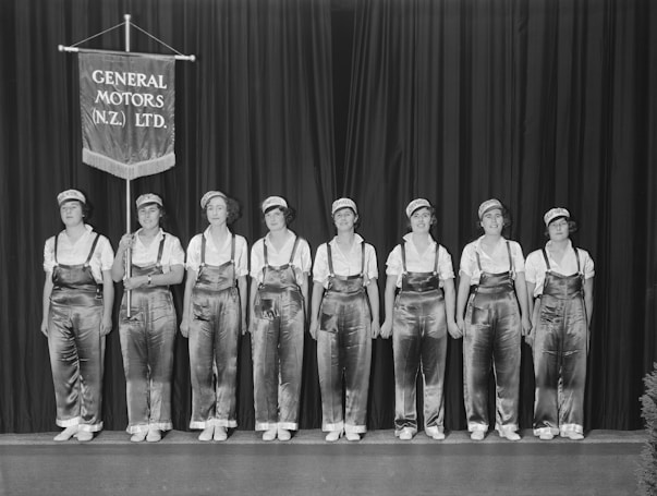 Seven women dressed in overalls and caps stand in a line against a backdrop. A banner with the inscription 'General Motors (N.Z.) Ltd.' is held by one of the women.