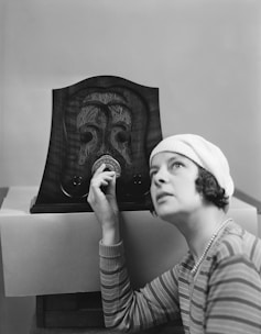 A woman in a striped shirt and cloche hat is adjusting the dial on an ornate vintage radio. The radio features intricate carved designs on its wooden cabinet.
