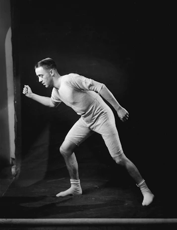 A person is wearing a vintage-style athletic outfit, composed of a form-fitting shirt and shorts, along with socks. The individual is posed in a running or athletic stance against a dark background.
