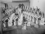 Multiple identical cardboard cutouts of a woman sitting on a wooden post, wearing a patterned sleeveless outfit, are arranged against a backdrop with soft lighting. Each cutout features the text 'Petone Quality Goods' and 'Newest Styles and Colourings.' The scene suggests a promotional or advertising setup.