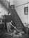 A woman is using a vintage vacuum cleaner on a leopard skin rug in a hallway with a staircase. The setting features ornate decorations including a patterned carpet on the stairs, framed pictures on the walls, a plant in a decorative pot, and antique furniture.