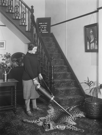 A woman is using a vintage vacuum cleaner on a leopard skin rug in a hallway with a staircase. The setting features ornate decorations including a patterned carpet on the stairs, framed pictures on the walls, a plant in a decorative pot, and antique furniture.