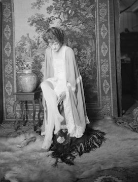 A woman in a vintage setting sits on a chair covered with a fringed cloth. She is wearing a robe with lace edges and adjusting her stockings. Behind her is an intricately designed tapestry depicting a pastoral scene with trees and buildings. On the left, a decorative vase rests on a small wooden table, and a pair of shoes is placed on a plush carpet on the floor.