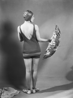 A person stands with their back to the camera, wearing a vintage swimsuit and a swim cap. They are holding an umbrella with a patterned design. The setting is minimalistic with a plain backdrop, suggesting an indoor studio environment.