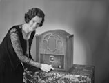 A woman is adjusting an ornate vintage wooden radio sitting on a table with a patterned cloth. She has short curled hair and is wearing a dark lace dress with a long necklace.