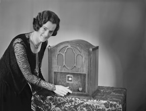 A woman is adjusting an ornate vintage wooden radio sitting on a table with a patterned cloth. She has short curled hair and is wearing a dark lace dress with a long necklace.