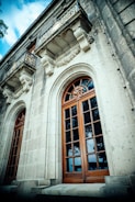 A detailed facade of a historic building with ornate stone carvings above large wooden-framed windows. Two small balconies with intricate wrought iron railings are visible, adding to the architectural elegance. The structure is made of light gray stone, creating a contrast with the rich brown wood of the windows.