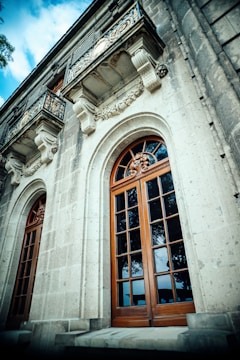 A detailed facade of a historic building with ornate stone carvings above large wooden-framed windows. Two small balconies with intricate wrought iron railings are visible, adding to the architectural elegance. The structure is made of light gray stone, creating a contrast with the rich brown wood of the windows.