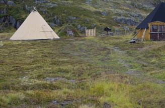 A large conical tent is set up on a grassy, slightly rocky landscape with a backdrop of hilly terrain. To the right, there are wooden structures, possibly small sheds or cabins, and a fenced area. The scene appears remote and natural.