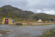 A rustic, wooden hut with a red door stands on gravel with a picnic bench nearby. There are two tents in the background, set against a backdrop of green, rocky hills under a cloudy sky. A wooden fence and a sign reading 'North Cape' are also visible.
