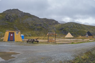 A rustic, wooden hut with a red door stands on gravel with a picnic bench nearby. There are two tents in the background, set against a backdrop of green, rocky hills under a cloudy sky. A wooden fence and a sign reading 'North Cape' are also visible.