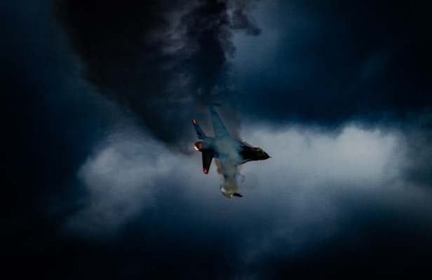 A fighter jet is soaring through a dramatically lit sky, surrounded by dark swirling clouds. The jet emits bright orange flames from its engines, creating a striking contrast against the ominous backdrop.