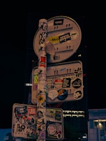 A street sign is densely covered with various stickers, including characters, logos, and texts in different languages and styles. These stickers range from simple text messages to complex graphics, completely obscuring the sign's original purpose.