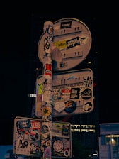 A street sign is densely covered with various stickers, including characters, logos, and texts in different languages and styles. These stickers range from simple text messages to complex graphics, completely obscuring the sign's original purpose.