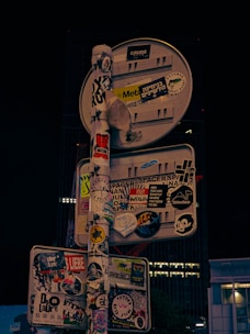 A street sign is densely covered with various stickers, including characters, logos, and texts in different languages and styles. These stickers range from simple text messages to complex graphics, completely obscuring the sign's original purpose.