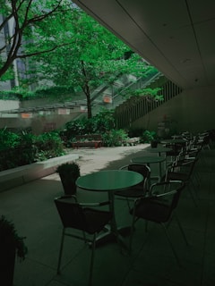 A peaceful indoor courtyard features green trees and plants, surrounded by a modern building. There are several empty tables and chairs, creating a serene setting for relaxation or conversation.