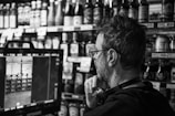 A person with glasses is attentively looking at a screen which is likely showing a camera feed in a store. Shelves filled with various bottles and items are visible in the background.