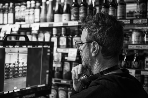 A person with glasses is attentively looking at a screen which is likely showing a camera feed in a store. Shelves filled with various bottles and items are visible in the background.