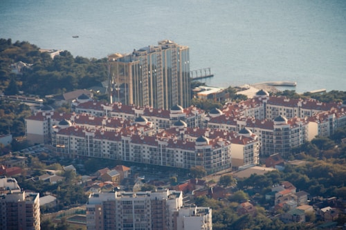 Aerial view of a coastal area with several residential buildings featuring red-tiled roofs and white walls. The structures are surrounded by dense greenery and situated near a large body of water. In the background, a taller building rises above the others, with a modern architectural style.