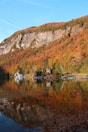 A serene lakeside scene with colorful autumn trees surrounding a calm body of water. Several cabins are nestled near the shore, reflecting beautifully in the lake. In the background, a rocky cliff rises against a clear blue sky, enhancing the natural, picturesque setting.