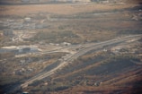 An elevated highway with multiple lanes curves through a hilly, semi-urban landscape. There is a prominent overpass, and nearby are several buildings, including a large white structure and residential houses. The surrounding area is dotted with trees and patches of grass, with a larger expanse of open land in the background.