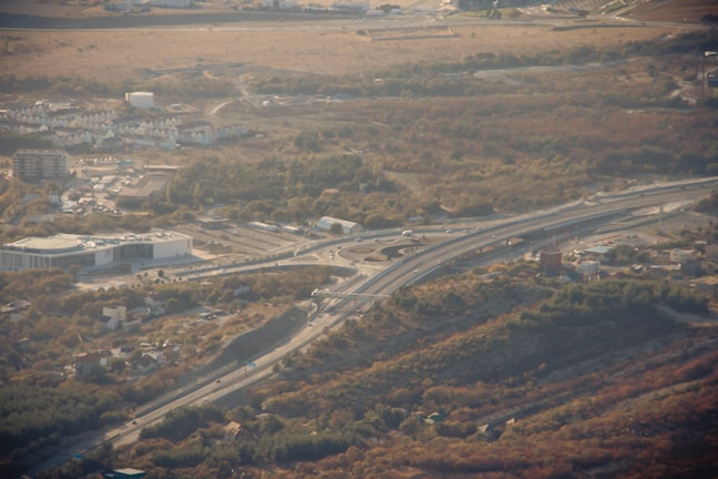 An elevated highway with multiple lanes curves through a hilly, semi-urban landscape. There is a prominent overpass, and nearby are several buildings, including a large white structure and residential houses. The surrounding area is dotted with trees and patches of grass, with a larger expanse of open land in the background.