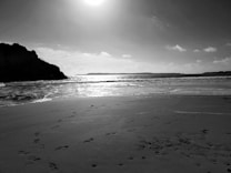 A serene black and white photograph of a beach with gentle waves lapping the shore. Footprints are visible in the sand, leading towards the water. The sun is high in the sky, casting a bright light over the ocean and creating a stunning contrast.
