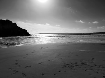 A serene black and white photograph of a beach with gentle waves lapping the shore. Footprints are visible in the sand, leading towards the water. The sun is high in the sky, casting a bright light over the ocean and creating a stunning contrast.