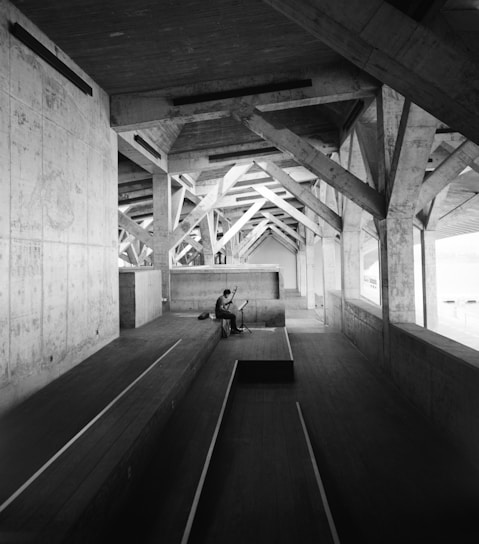 A person is seated on a wooden platform inside a large, industrial-style structure with exposed beams and concrete walls. The environment appears vast and empty except for the lone individual playing a musical instrument. The architecture features a series of triangular trusses that create an interesting pattern of light and shadows.