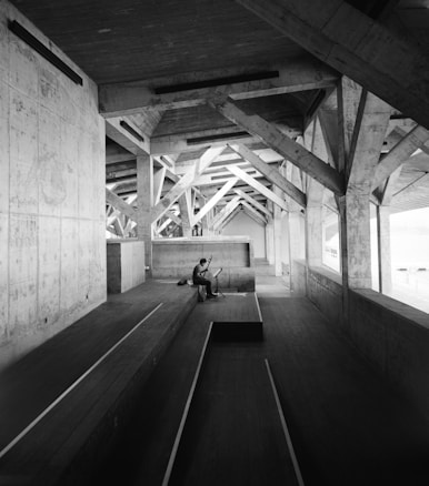A person is seated on a wooden platform inside a large, industrial-style structure with exposed beams and concrete walls. The environment appears vast and empty except for the lone individual playing a musical instrument. The architecture features a series of triangular trusses that create an interesting pattern of light and shadows.