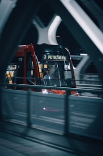 A red tram moving along a bridge at night. The tram has the number 3 and the destination 'PETRZALKA' displayed on its front. There are several passengers inside, visible through the windows. The bridge has a modern architectural design with white curved supports.