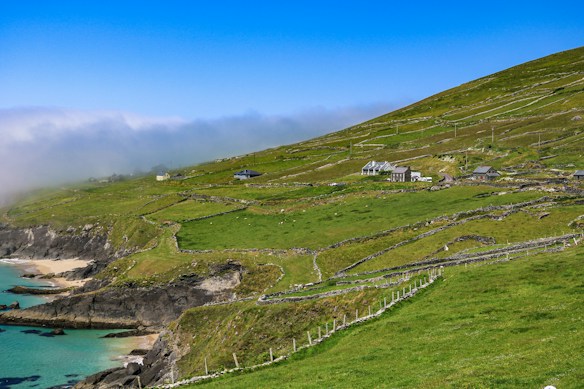 A scenic coastal landscape featuring lush green hills with scattered houses and stone fences. The foreground showcases rocky cliffs leading down to a turquoise sea and sandy beach. Mist partially covers the horizon, adding an ethereal quality to the scene.