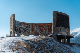 A large, curved monument made of stone and concrete is adorned with colorful murals depicting various figures and abstract designs. It is situated on a snow-covered hillside with a clear blue sky overhead. In the background, snow-capped mountains are visible, adding to the scenic setting.